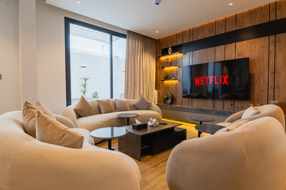 The living area features a modern design with two plush, cream-colored sofas arranged around a low black coffee table. A large television screen displays the Netflix logo. Natural light flows through a sizable window, complemented by soft drapes.