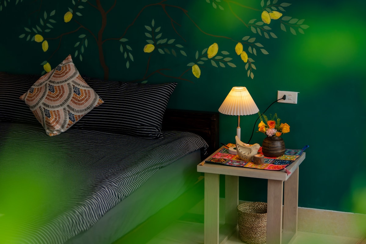 A cozy bedroom features a king-size bed with dark striped bedding against a vibrant green wall adorned with hand-painted foliage. A small lamp illuminates a bedside table, which holds a decorative bowl and a bouquet of flowers, adding a touch of warmth to the space.