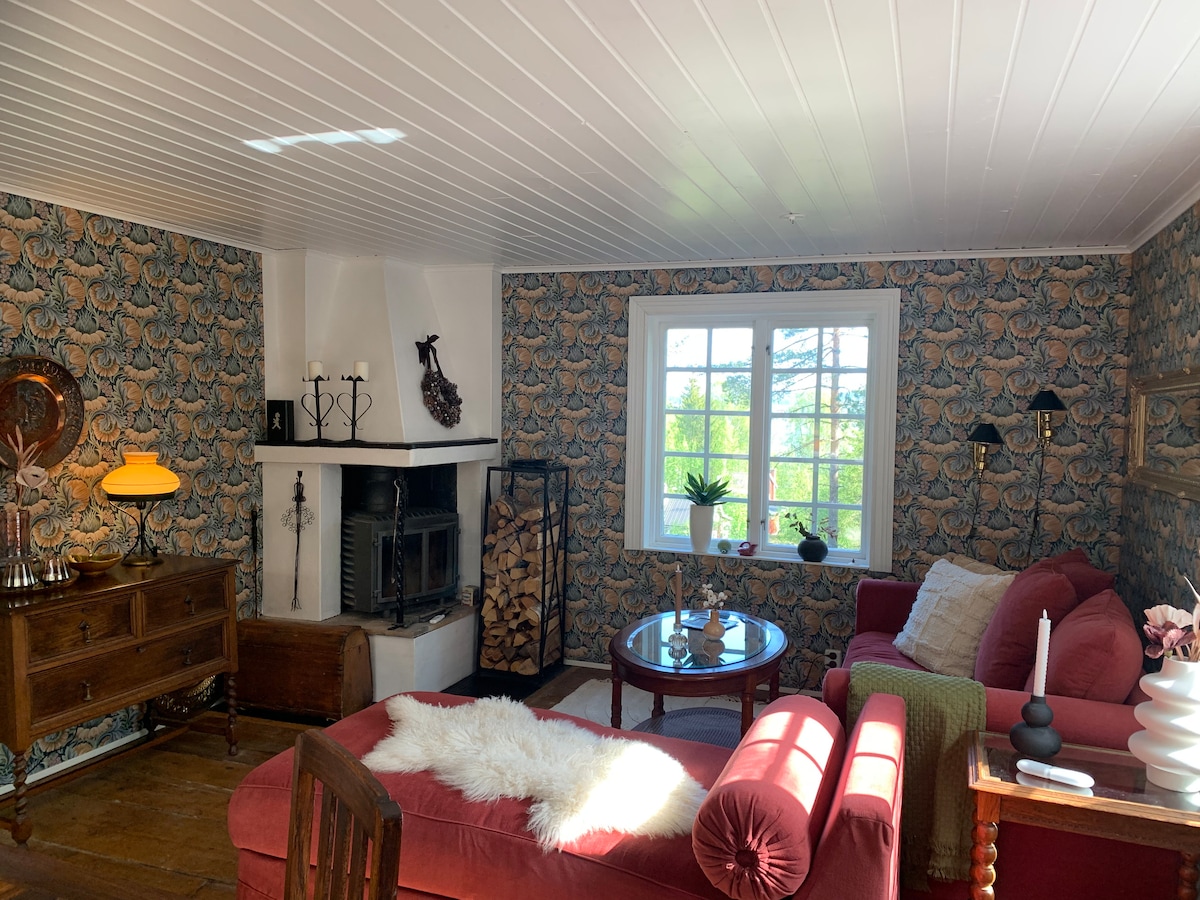 A welcoming living area features patterned wallpaper and a cozy red couch. A round coffee table sits at the center, with a small lamp providing light. A wood-burning stove is positioned next to neatly stacked logs, and a large window offers views of the surrounding nature.