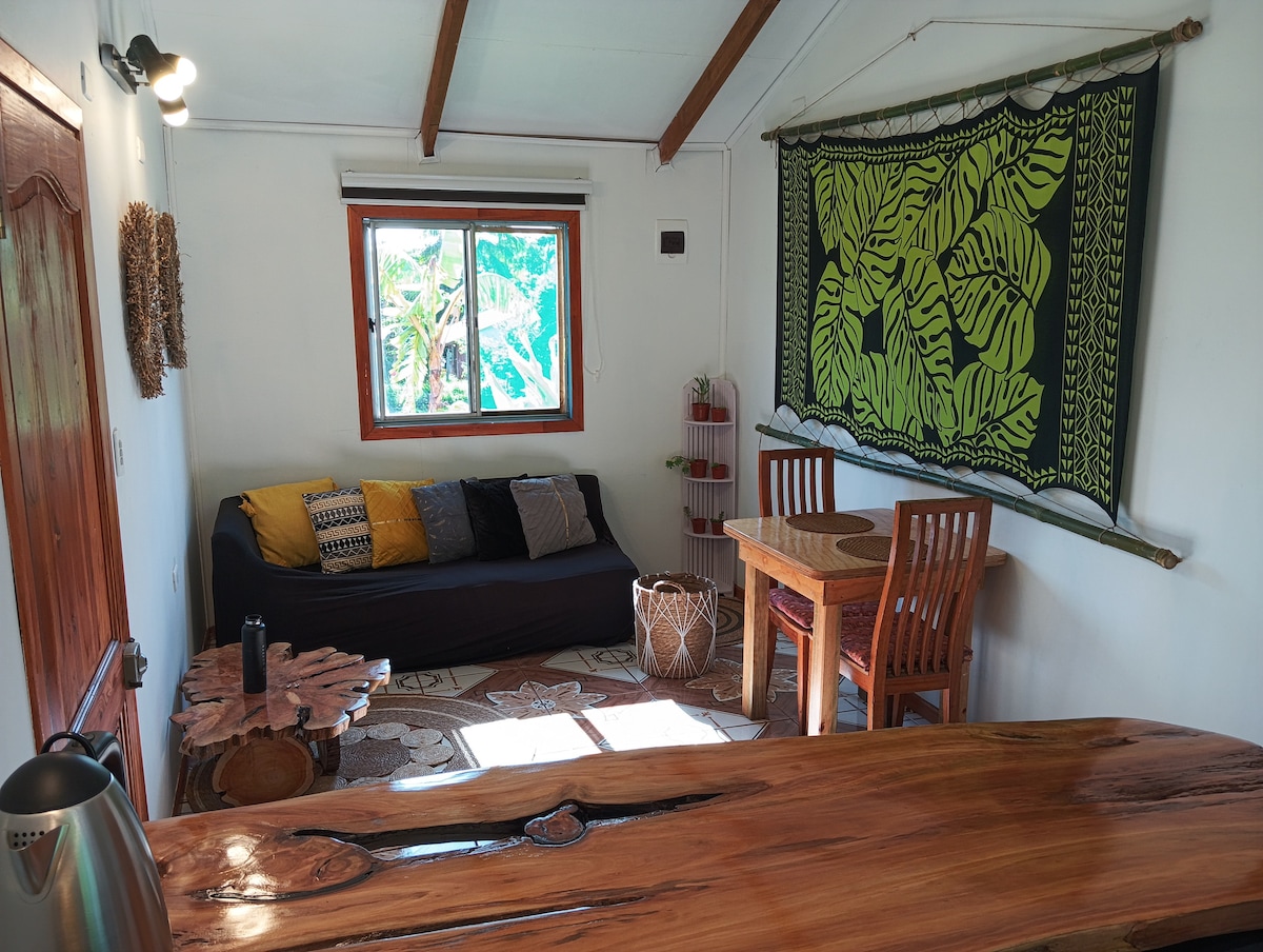 An inviting interior space features a dark sofa with colorful cushions and a natural wood dining table surrounded by four chairs. A striking wall hanging with green patterns adds a vibrant touch, while a large window offers views of the garden.