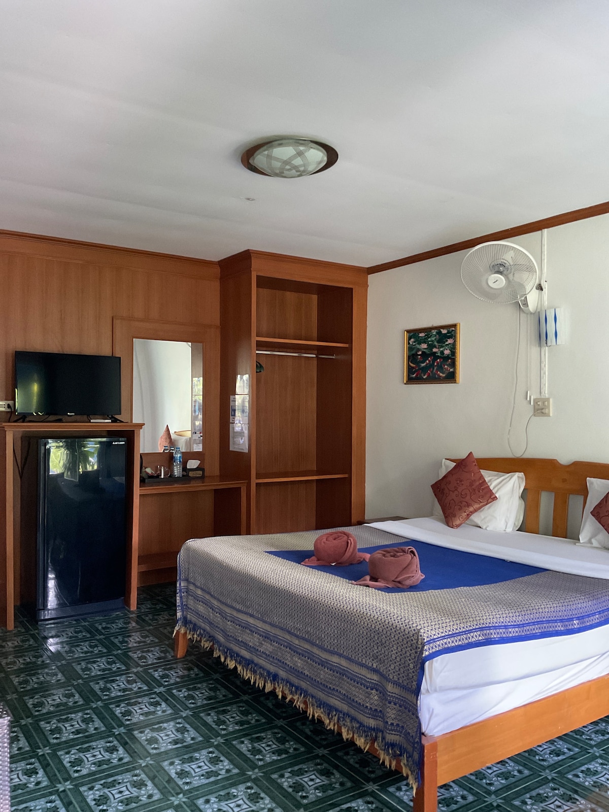 A simple guest room is furnished with a spacious bed, small table, and a television. A mirrored wardrobe and mini-fridge are included. Decorative pillows are placed on the bed. Natural light enters through a window, while a ceiling fan provides airflow.