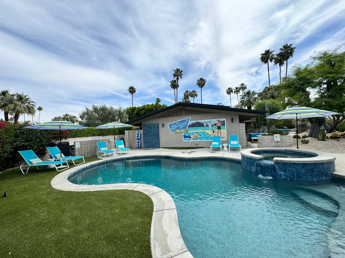 A vibrant outdoor space features a saltwater pool surrounded by lush landscaping. Light blue loungers and umbrellas provide relaxation options, while a stone spa is set beside the pool. Palm trees rise in the background, complementing the serene atmosphere.