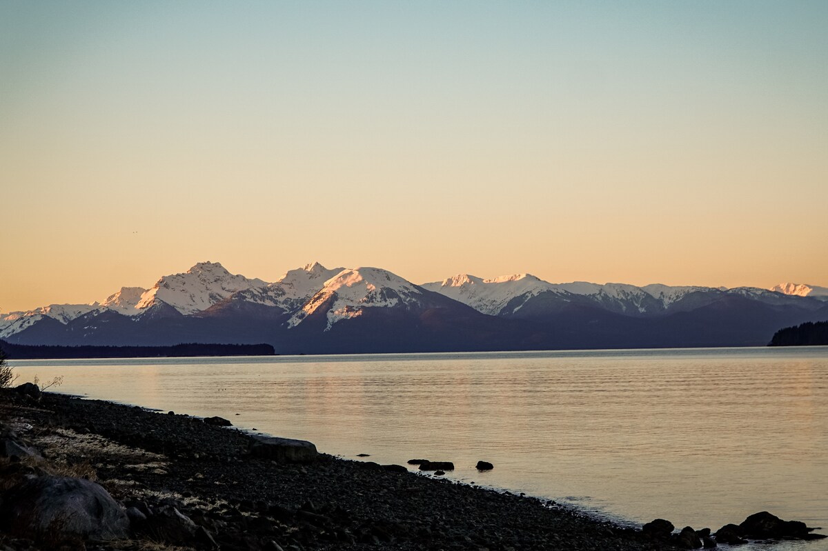 Snow-capped peaks of the Chilkat Mountains are visible against a soft sunset sky. The tranquil waters of the insides passage reflect the colors of nature, while a rocky shoreline is seen in the foreground, completing this serene coastal landscape.