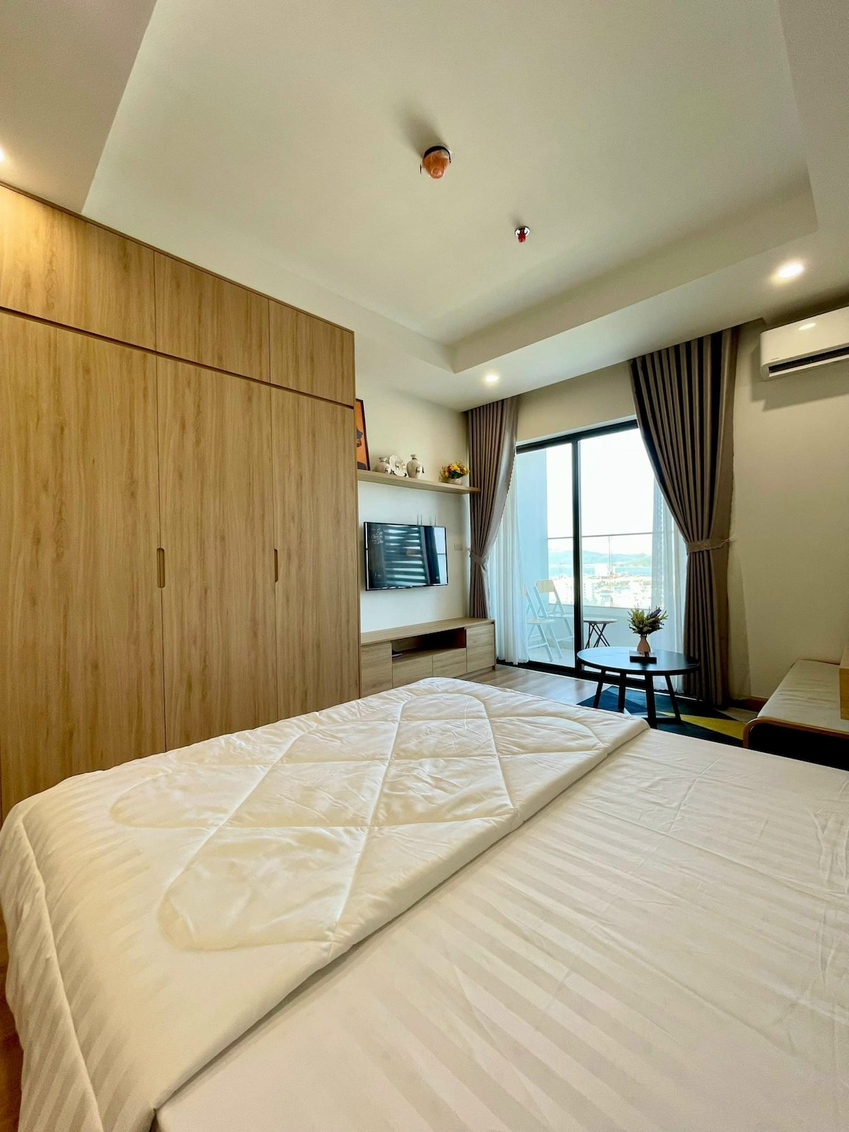 A cozy bedroom features a spacious bed covered with a light quilt. Wooden wardrobes provide ample storage. A balcony door allows natural light to enter, revealing a small outdoor seating area with two chairs. A wall-mounted television is visible, enhancing comfort and convenience.