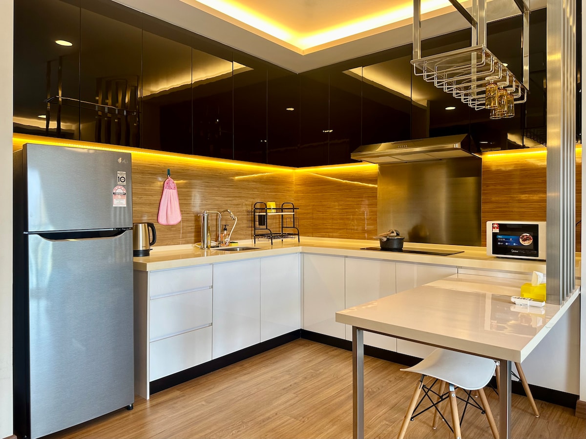 A modern kitchen features sleek black cabinets and warm wood accents. Stainless steel appliances, including a refrigerator and a stovetop, create a functional space. A small dining table is visible, accompanied by minimalist white chairs. Soft lighting enhances the contemporary design.