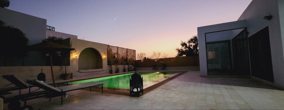 An outdoor area features a private pool illuminated in green water, surrounded by soft tile flooring. A shaded lounge area is equipped with sun loungers, while decorative lanterns cast a gentle glow. The sky transitions to twilight, showcasing soft hues of purple and orange.