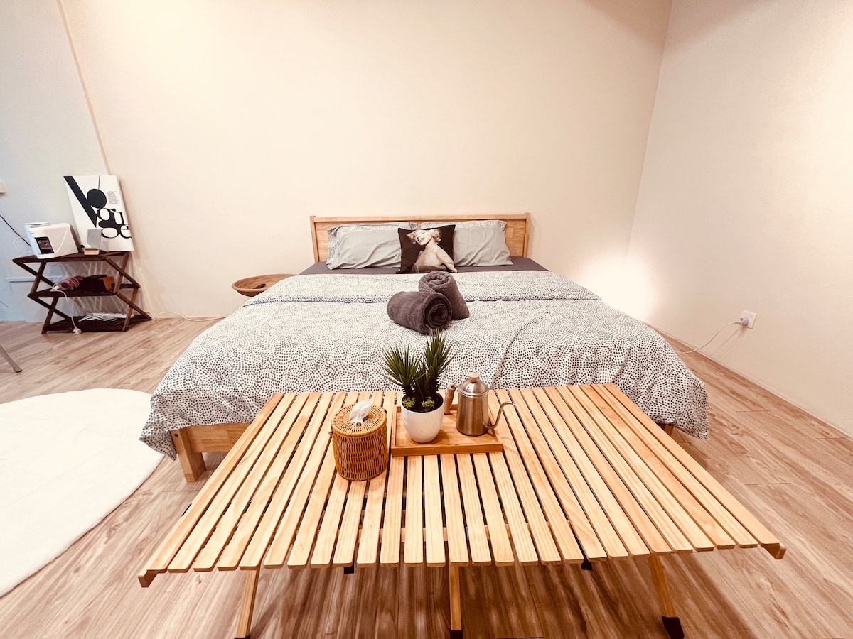 A spacious sleeping area features a simple bed with neutral-toned linens and a decorative pillow. A rolled towel is placed on top. A wooden, slatted table holds a small plant and serving items, complemented by natural lighting from the space.