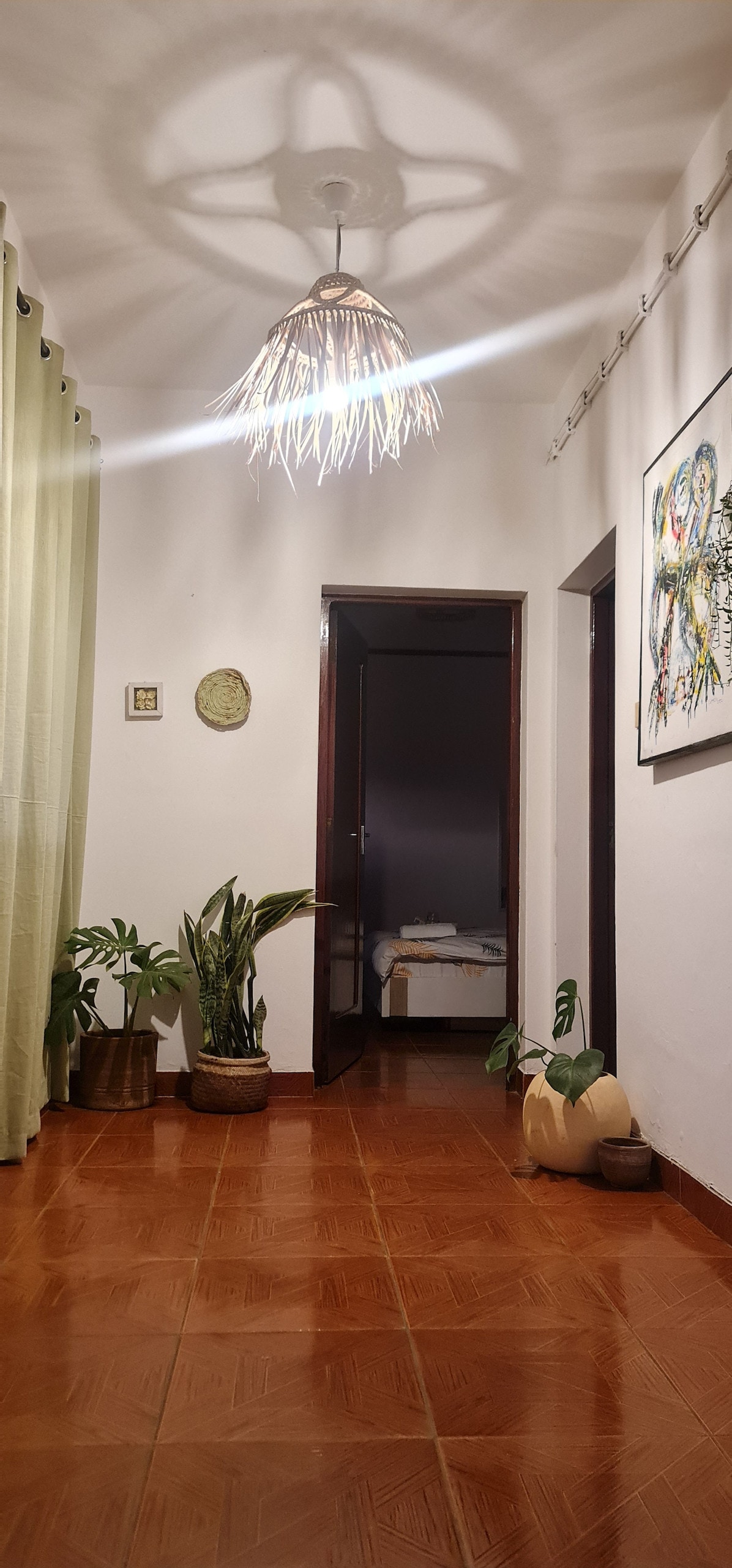 A hallway is featured with warm-toned tile flooring, leading to a softly lit room at the end. A modern hanging light fixture casts intricate shadows on the wall. Green curtains frame the doorway, while potted plants add a touch of nature along the sides.