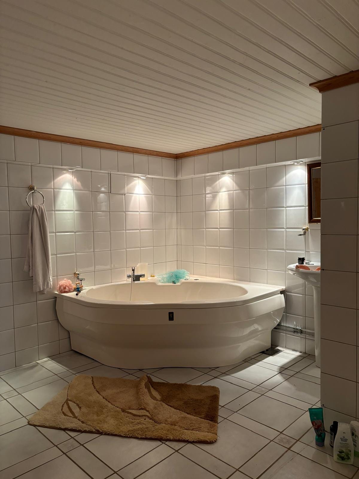 A rounded white bathtub is situated in a well-lit bathroom, surrounded by glossy white tiles. Soft lighting from above highlights the contours of the tub. A beige mat lies on the tiled floor, complemented by a towel hanging nearby and various toiletries arranged on a shelf.