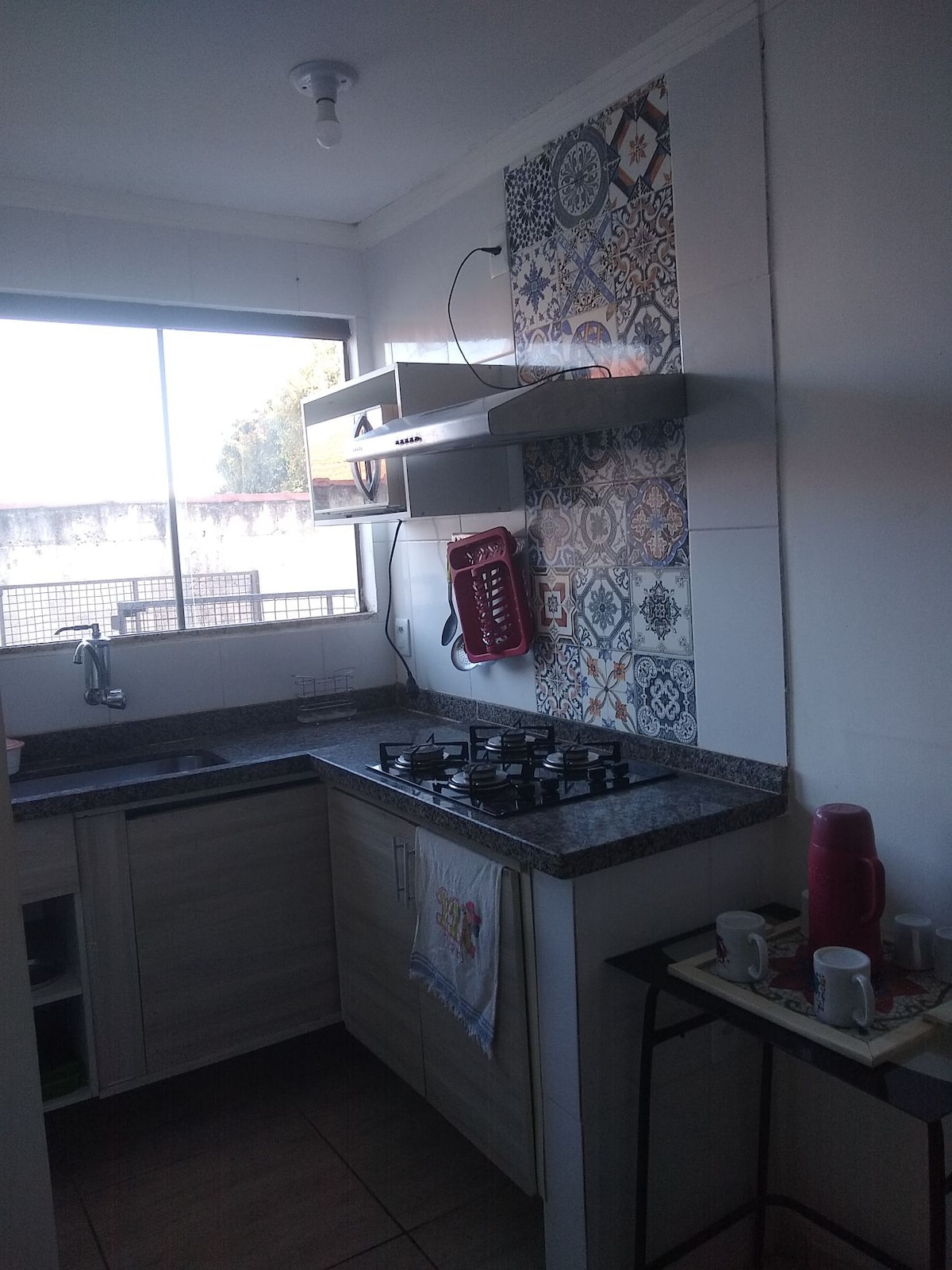 A functional kitchen space features light wooden cabinetry and a granite countertop. A gas stove is positioned beneath an exhaust hood. Colorful tiled accents provide a decorative touch. A window allows natural light, with a small table set beside it, displaying kitchen items.