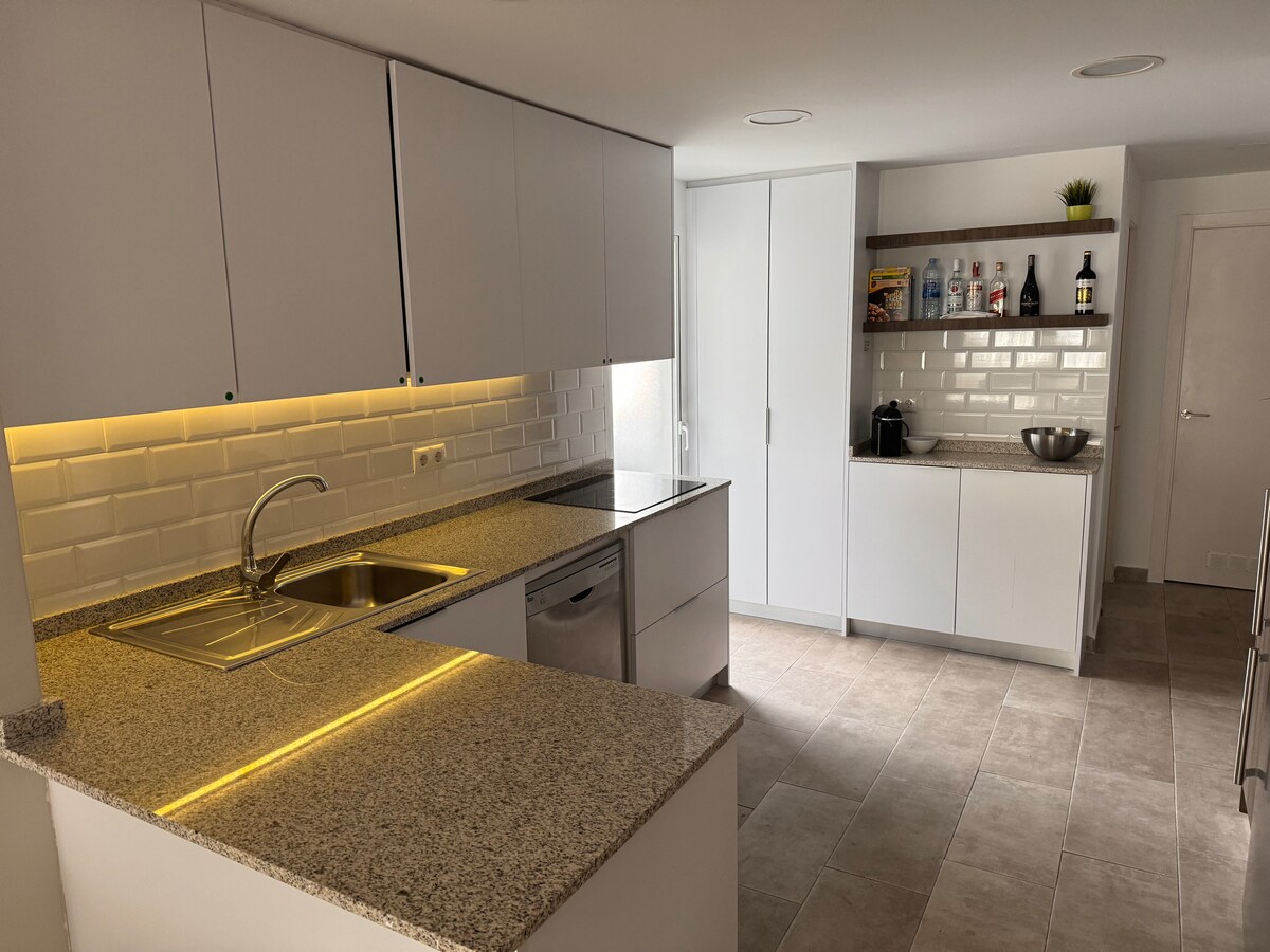 A modern kitchen features sleek white cabinetry with minimalist handles and a granite countertop. A stainless-steel sink and dishwasher are visible beneath warm under-cabinet lighting. Open shelving displays various bottles and a bowl, adding functional elements to the design.