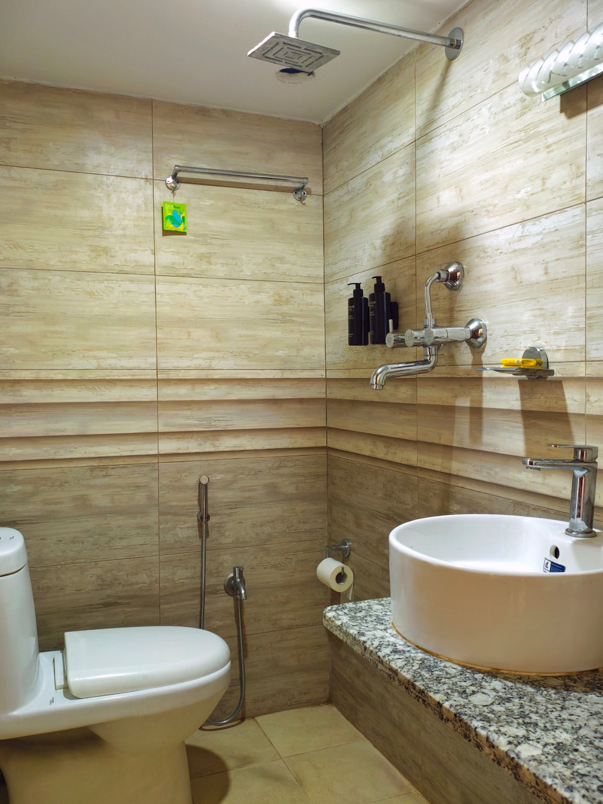 An attached bathroom is depicted, featuring a wall-mounted sink set atop a granite countertop. A modern toilet and stainless steel shower fixtures are visible. Neutral-toned tiled walls create a clean and streamlined appearance, while toiletries are arranged on a shelf for convenience.