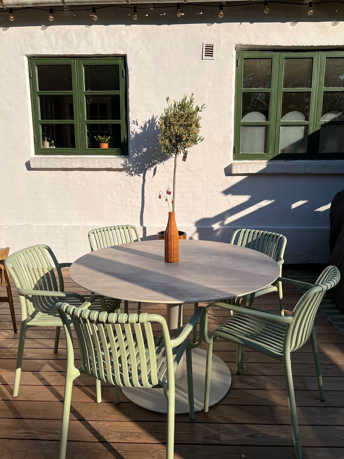 An outdoor dining area features a round table surrounded by six green chairs. A single amber vase with an olive branch is placed at the center of the table. Sunlight casts soft shadows on the wooden deck and white wall behind, highlighting the window's green frames.