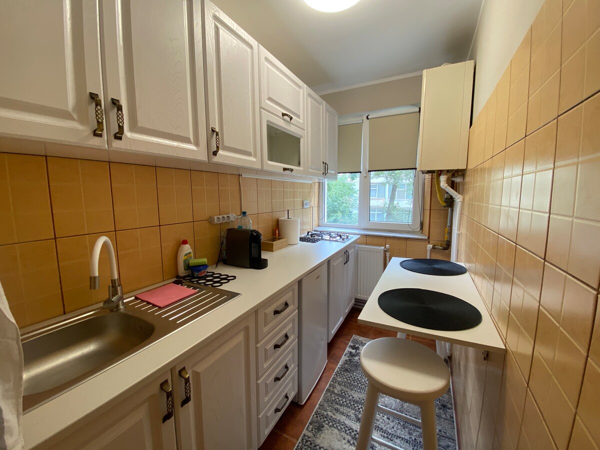 A bright kitchen is showcased, featuring white cabinetry and warm-colored tiled walls. A streamlined counter includes a sink and coffee maker, with a small dining area illuminated by natural light. Two minimalist stools are positioned at a compact table against the wall.