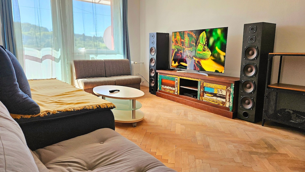 A cozy living area features a comfortable sofa and an armchair positioned near a low coffee table. A large television is mounted on a colorful cabinet, complemented by tall speakers on either side. Light streams through the windows, enhancing the room's calm ambiance.