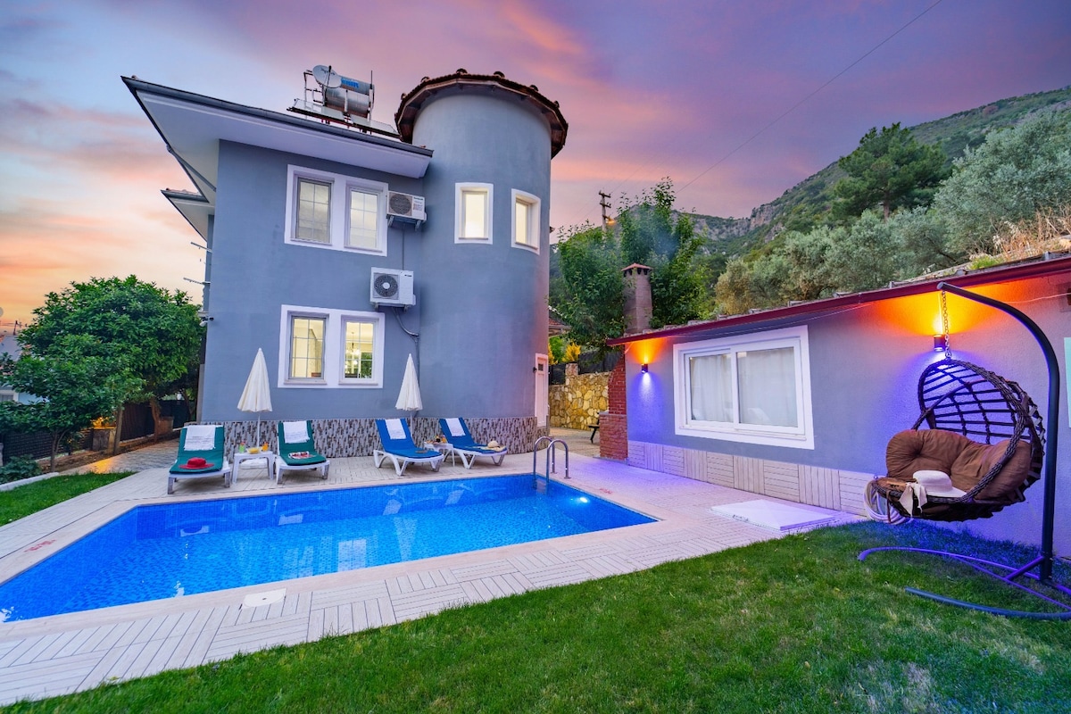 A two-story villa is shown, featuring a round turret and exterior walls painted in a soft grey hue. The well-maintained garden includes a refreshing swimming pool, lounge chairs, and a cozy hanging chair, all illuminated by warm outdoor lighting at dusk.