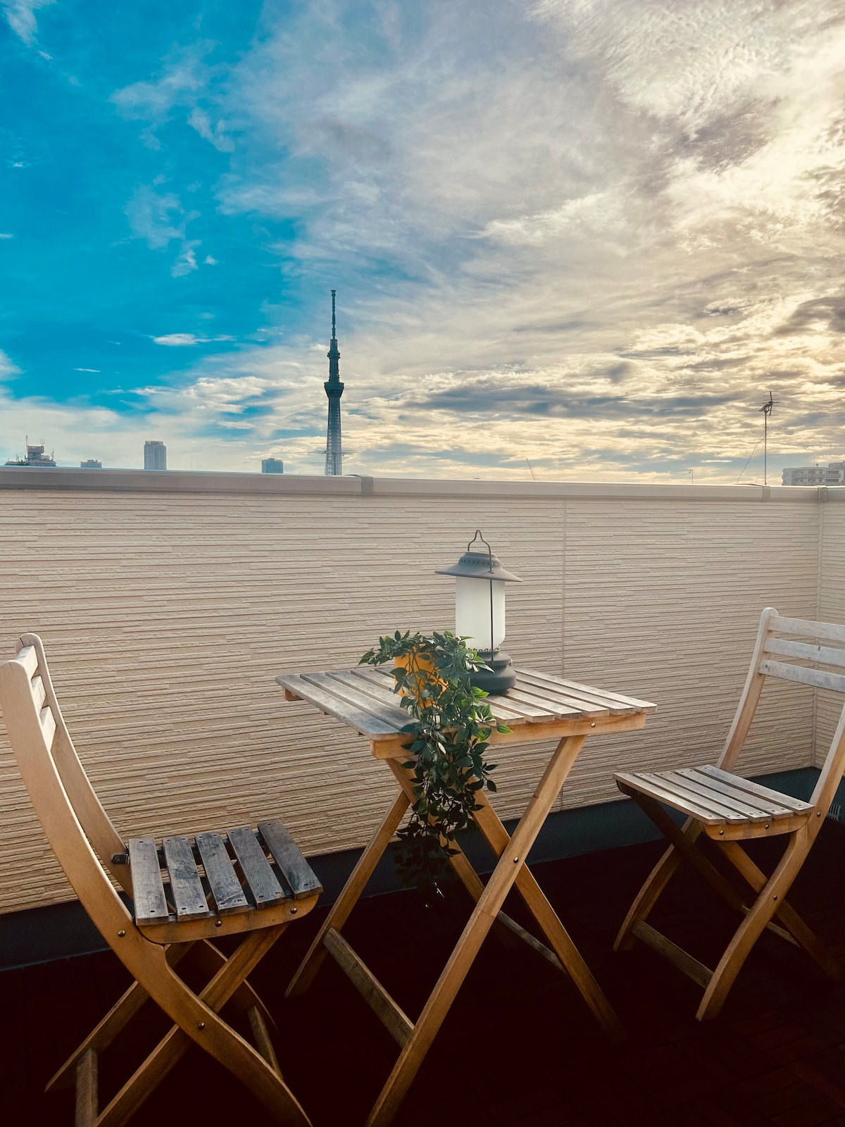 A charming rooftop space features a small wooden table and two folding chairs, inviting relaxation. A lantern is positioned on the table amidst greenery. The Sky Tree is visible in the distance, set against a backdrop of dynamic clouds and a softly lit sky.