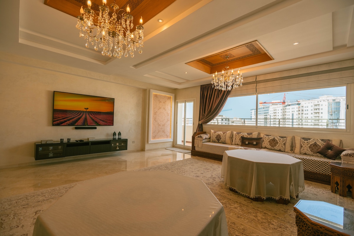 A spacious living area features a large flat-screen TV mounted on the wall, complemented by an elegant chandelier. Natural light streams through expansive windows adorned with curtains. Comfortable seating is arranged around a decorative coffee table, with intricate patterns visible on the furnishings.