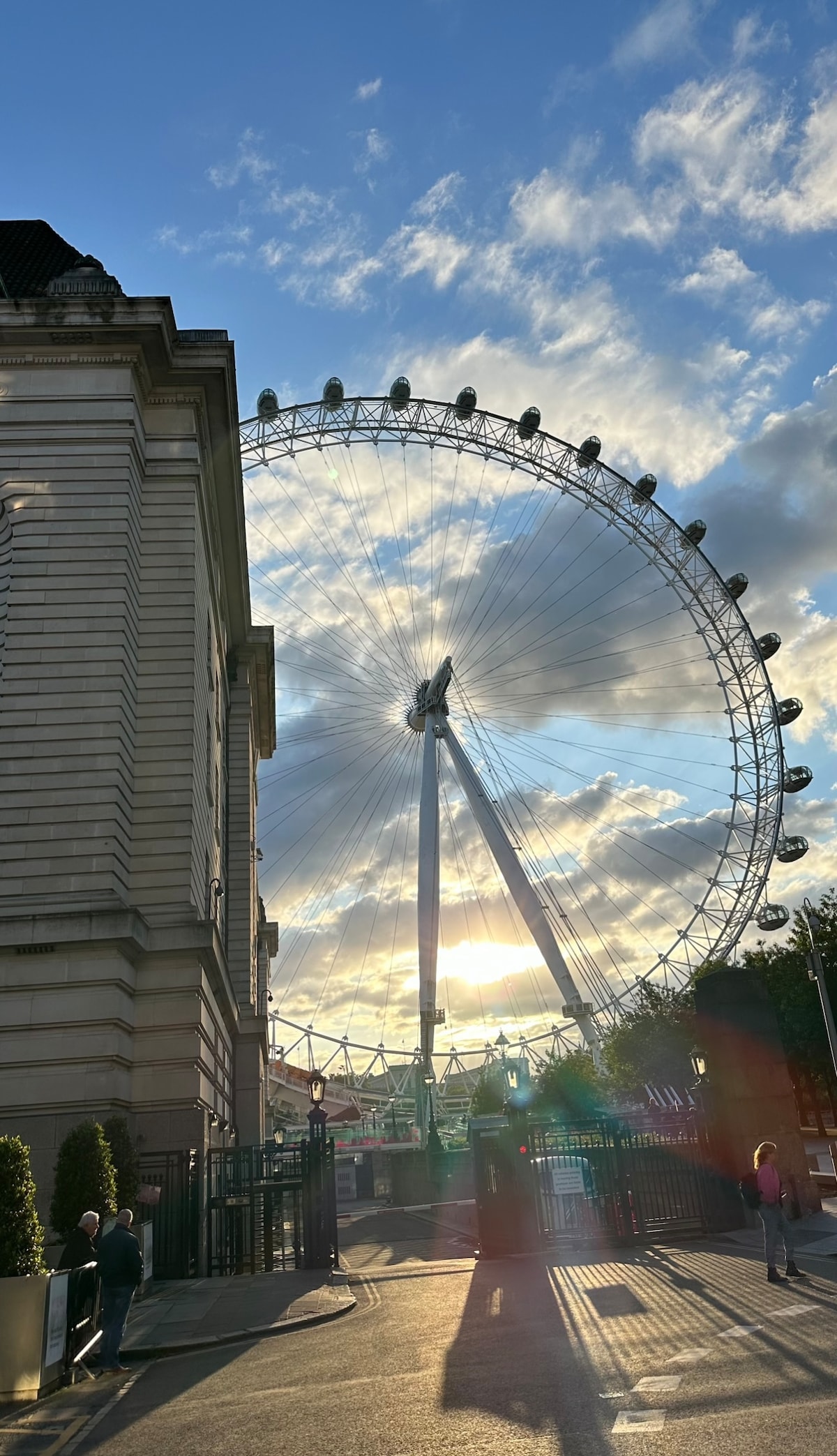 The London Eye is prominently featured in this image, partially obscured by a building. The large, rotating structure contrasts against a cloudy sky, with sunlight peeking through, creating an atmospheric backdrop. The entrance area includes greenery and hints of visitors approaching.