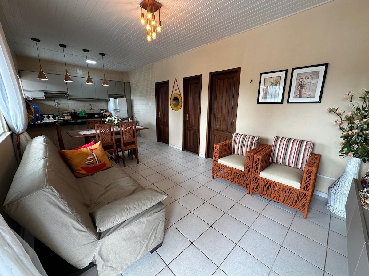 The living area features a light-colored sofa bed and two wicker chairs with striped cushions. A wooden dining table is positioned nearby, complemented by pendant lighting. The space is brightened by natural light and showcases a warm, inviting layout with doors leading to additional rooms.