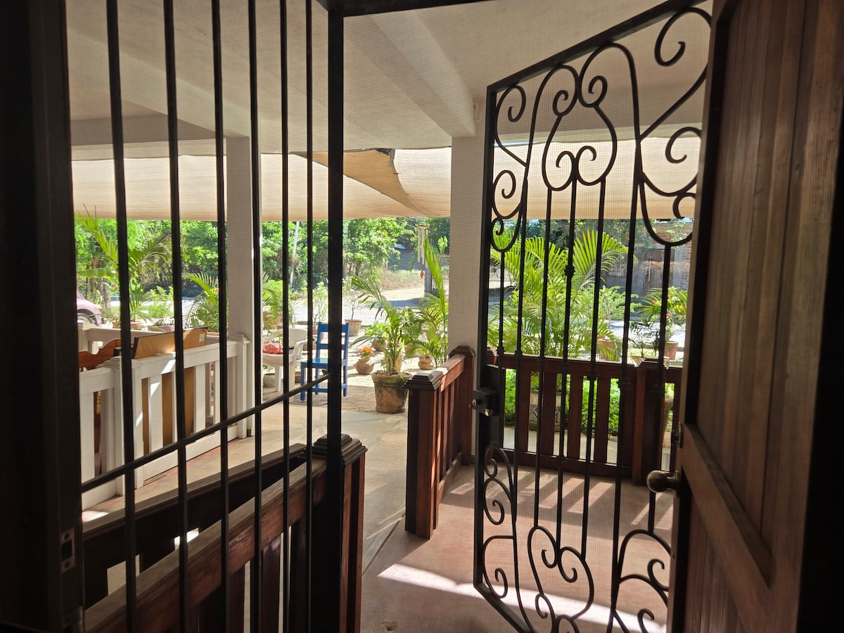 A view through an ornate iron gate reveals a spacious outdoor area filled with greenery. The entrance features a wooden door, leading to a patio with various seating options and natural light filtered through a canopy above.