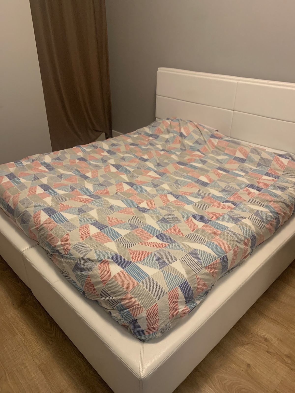 A spacious bed is dressed with a patterned comforter featuring geometric shapes in soft pastel tones. The headboard is sleek and white, while light wood flooring adds warmth to the area. A window with flowing brown curtains is visible in the background.