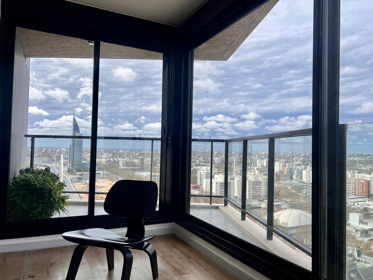 A corner of the apartment features large glass windows offering panoramic views of the urban landscape. A sleek black chair is positioned near the windows, with a potted plant adding a touch of greenery to the space. The sky is dotted with clouds.