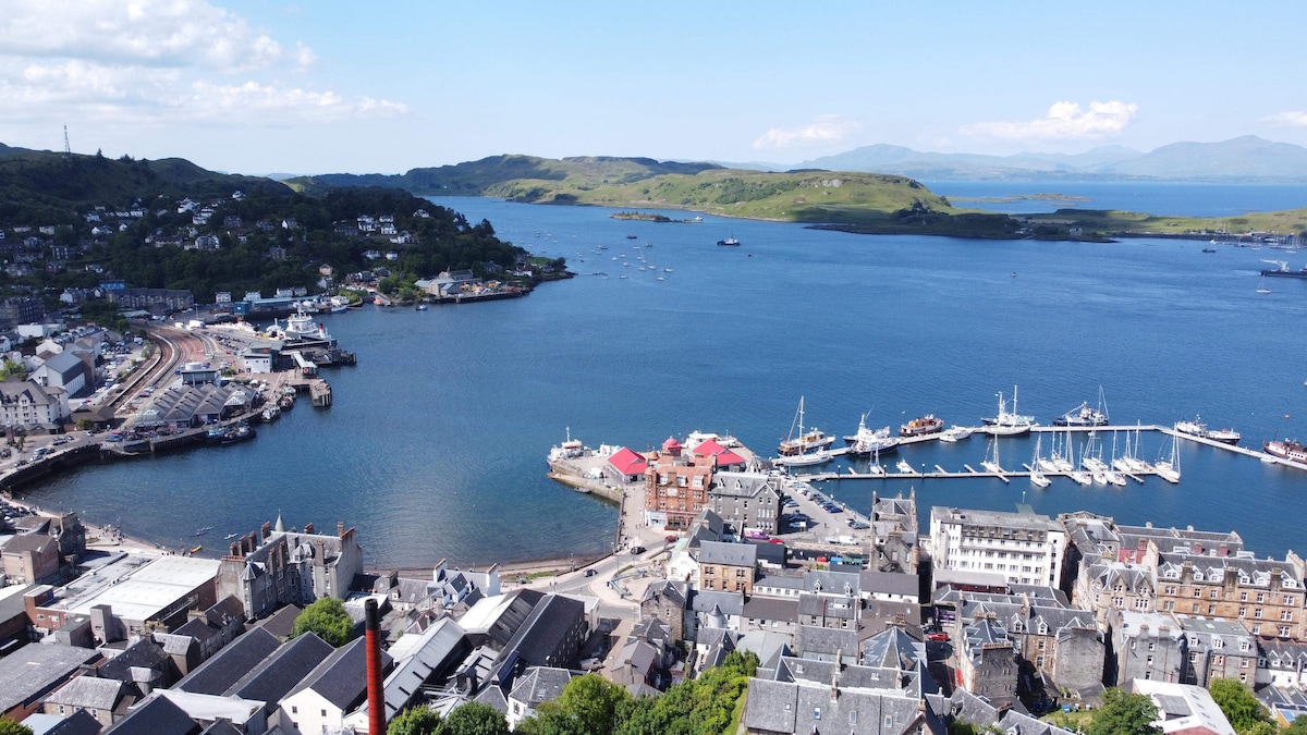 A panoramic view of a coastal town is displayed, featuring a vibrant harbor with boats anchored and a mixture of historical buildings lining the waterfront. Lush hills rise in the background, and the expansive blue water is dotted with small islands.