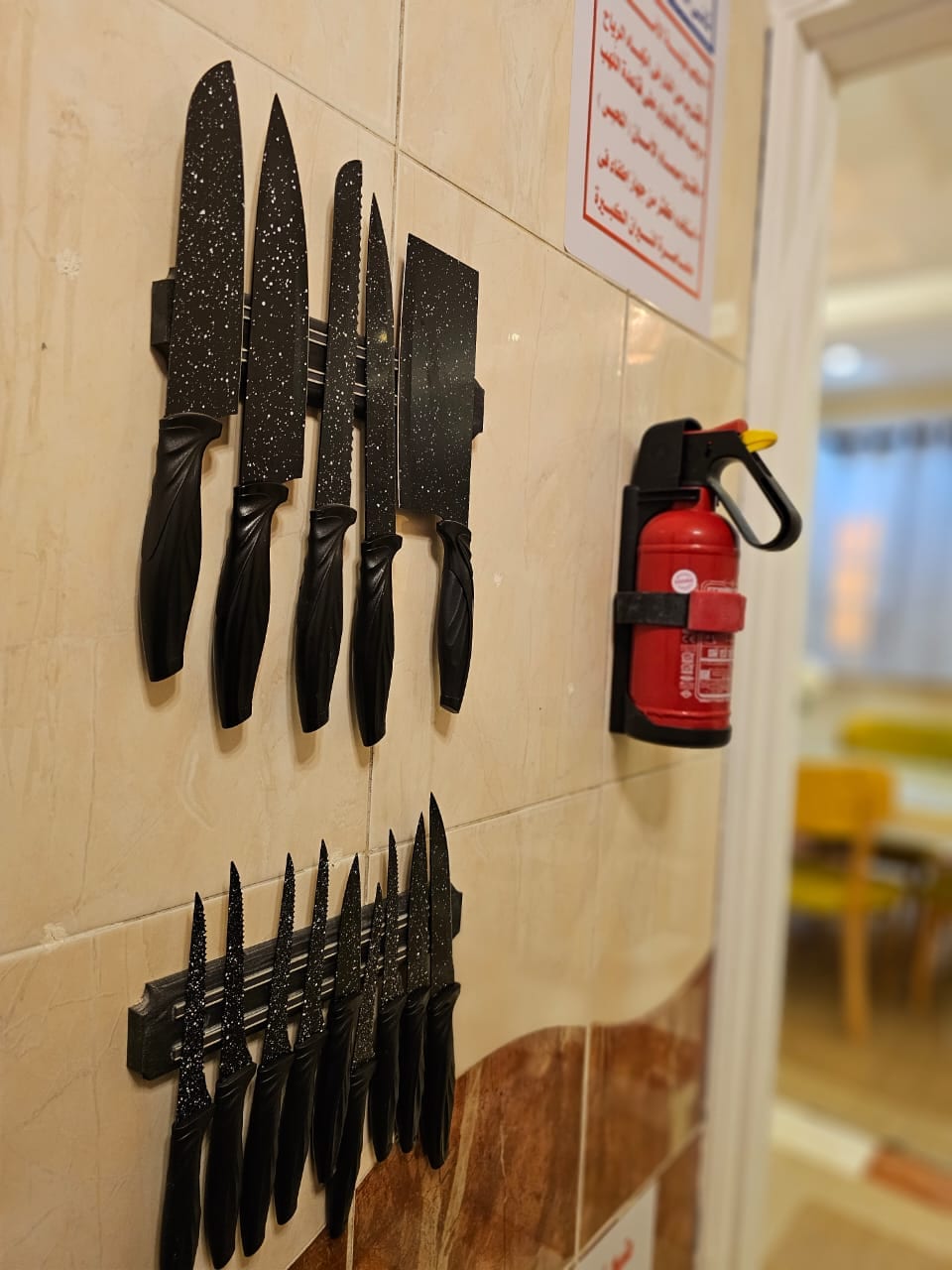 A wall-mounted knife holder displays an assortment of kitchen knives, featuring various sizes and blade styles. Adjacent to the knives, a fire extinguisher is securely mounted, ensuring safety in the kitchen area. The background shows a glimpse of a dining space with warm lighting.