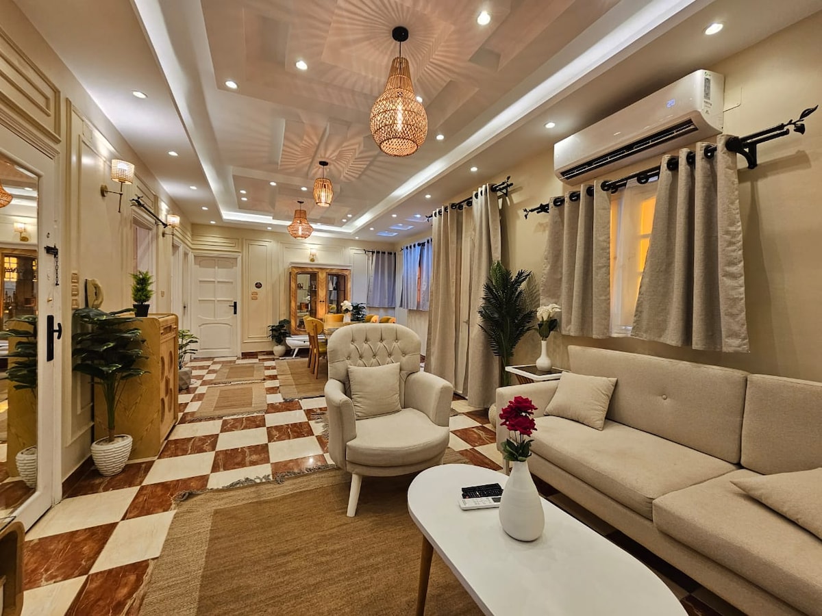 The inviting living area features a light-colored sofa and an armchair, complemented by a central coffee table. Soft lighting from decorative fixtures and wall sconces enhances the warm ambiance, while large windows with curtains allow natural light to filter in. A patterned area rug adds texture to the tile floor.