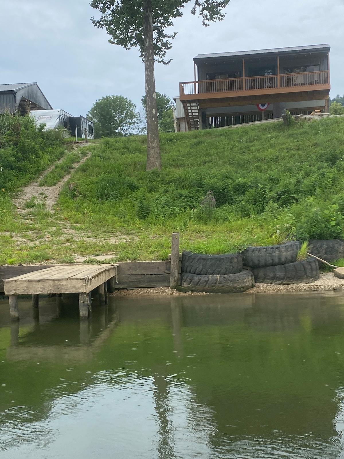 A two-level cabin is situated on a grassy slope overlooking a calm body of water. A wooden dock extends into the water, accompanied by tire barriers. The covered deck of the cabin offers views of the surrounding greenery and waterway.