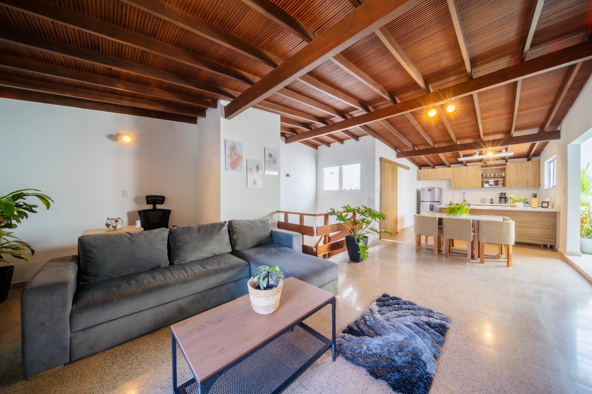 An open living area is illuminated by soft lighting, featuring a comfortable dark sofa and a wooden coffee table. Potted plants add natural elements to the space. The kitchen area is visible in the background, with modern appliances and a dining island, enhancing the room's practicality.