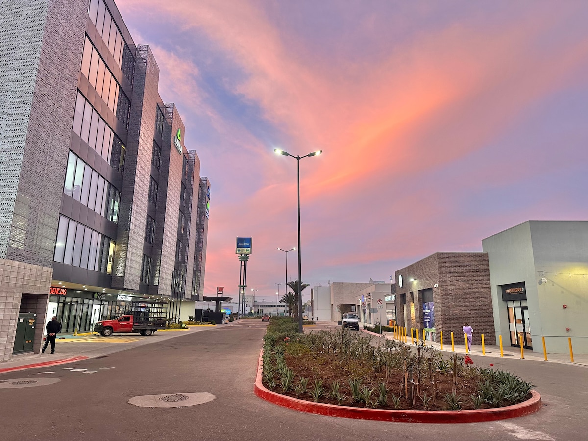 An urban landscape is presented at dusk, showcasing modern buildings under a sky painted with soft pink hues. Pathways are lined with plants, and streetlights illuminate the area, inviting evening strolls.