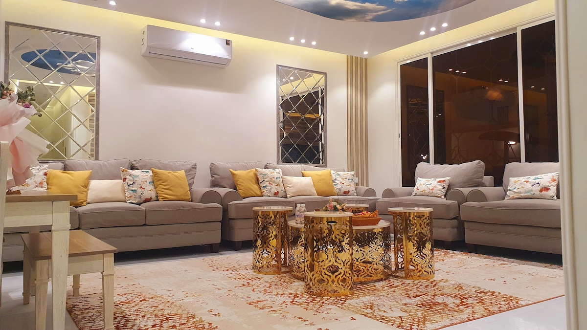 A spacious living area features several light grey sofas adorned with cushions in soft tones. Two intricately designed coffee tables sit on a large area rug, adding warmth to the space. Large windows offer views of the outside, while ambient lighting enhances the room's comfort.
