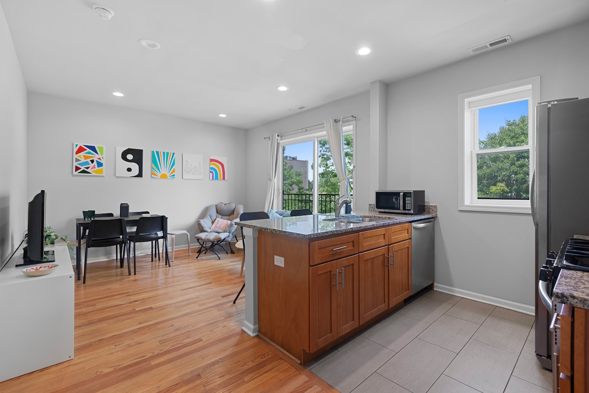 An open living area features a spacious kitchen with wooden cabinets and stainless steel appliances. A dining table is set for six, while modern artwork adorns the walls. Natural light streams in through large windows, enhancing the inviting atmosphere of the space.