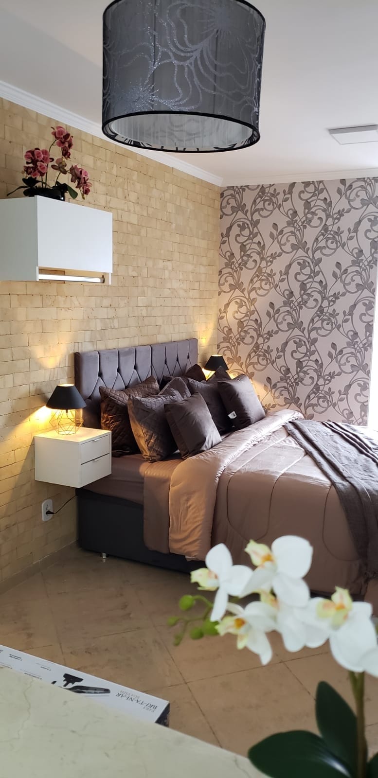 The bedroom features a comfortable Queen bed with an upholstered headboard, complemented by decorative pillows and a textured blanket. A stylish wallpaper and a brick accent wall enhance the decor. Soft lighting is provided by wall sconces, creating a cozy ambiance.