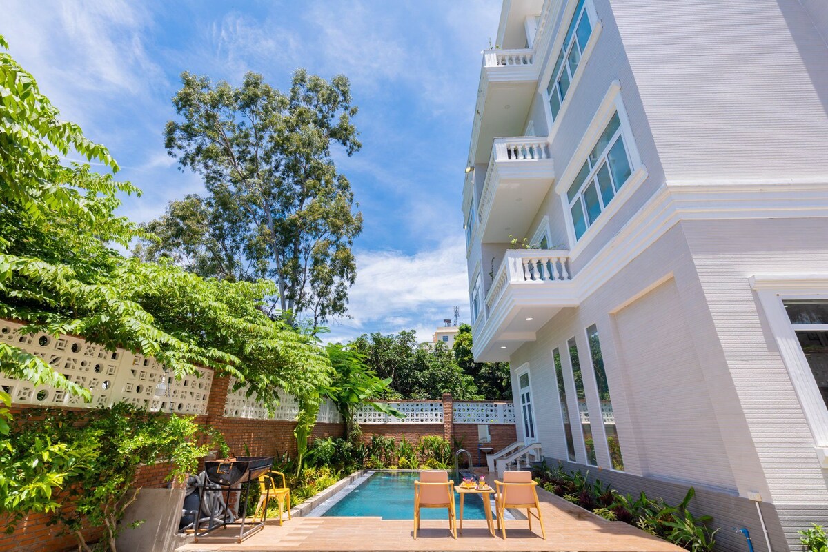 The villa features a spacious outdoor area with a clear pool surrounded by greenery. Two lounge chairs face the water, providing a relaxing space under the sun. The light-coloured exterior of the villa contrasts with the vibrant foliage, creating a serene atmosphere.