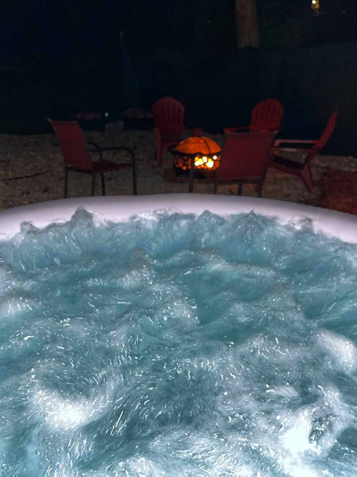 A bubbling hot tub filled with clear blue water occupies the foreground, while a fire pit surrounded by red chairs is seen in the background. The warm glow of the fire contrasts with the dark surroundings, creating a cozy outdoor ambiance.