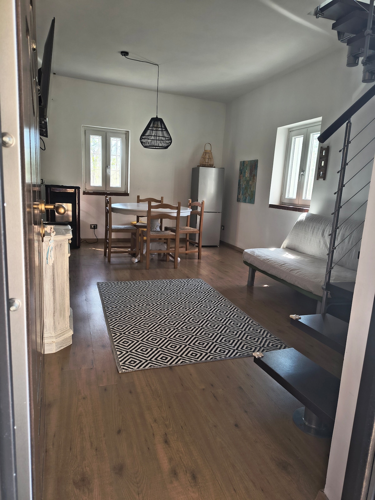 A cozy living space is presented with a sliding sofa bed and a dining area featuring a wooden table surrounded by four chairs. Natural light enters through two windows, complemented by a black pendant light hanging above. A patterned rug lies on the wooden floor, enhancing the environment.