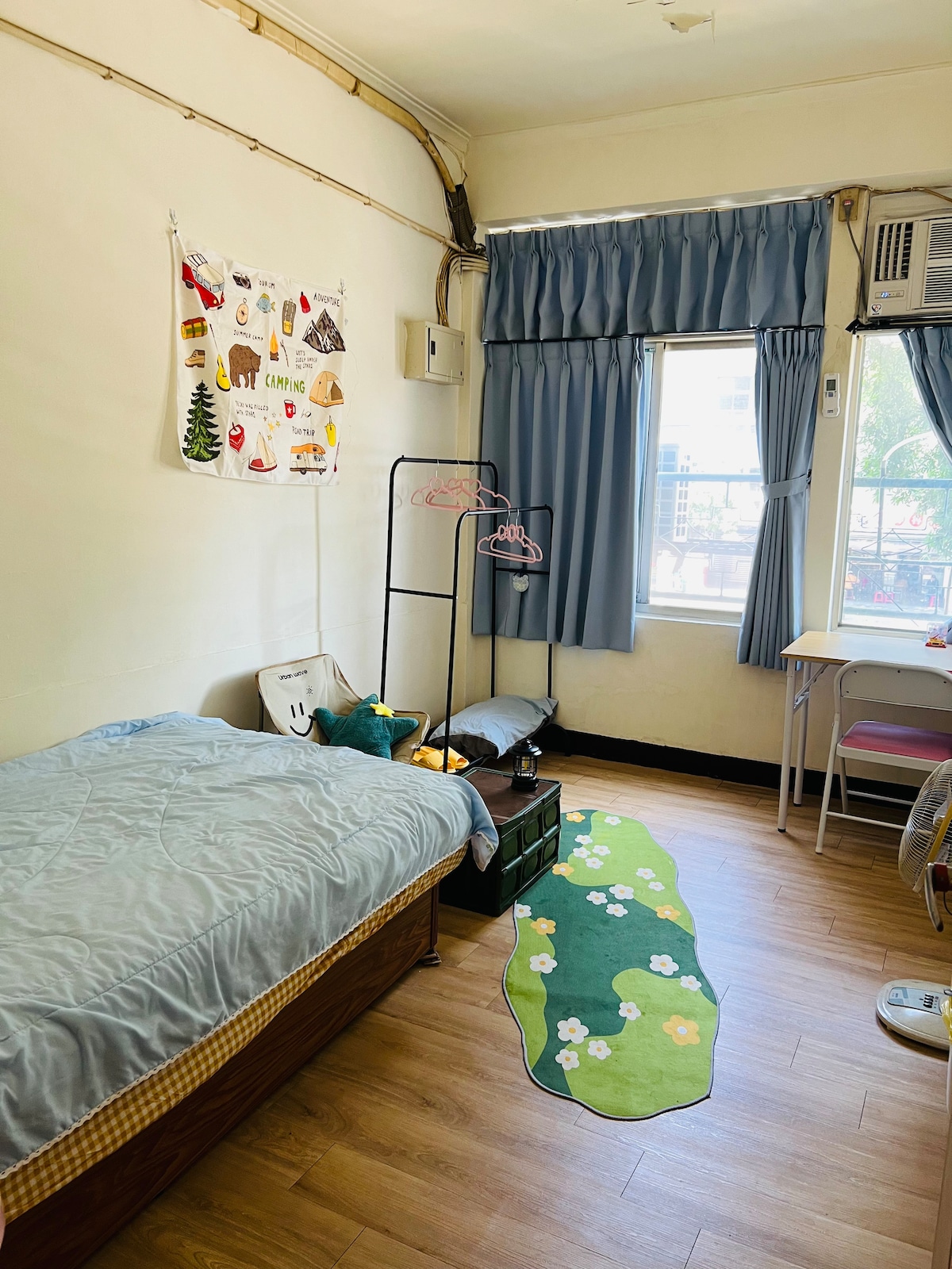 A serene room features a cozy bed with a light quilt, accompanied by a rug with floral patterns. Natural light enters through a window, illuminating the space. A clothing rack and a desk are present, adding functionality to the tidy environment.