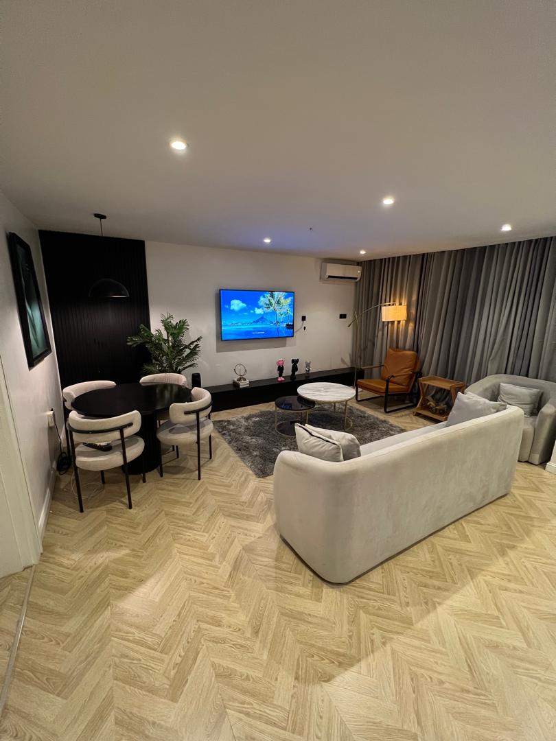 A modern living area is illustrated, featuring a soft grey sectional sofa and two stylish armchairs surrounding a small coffee table. A well-lit space is enhanced by a large artwork and a flat-screen TV, with a floor lamp providing additional lighting. A muted rug underfoot ties the area together.
