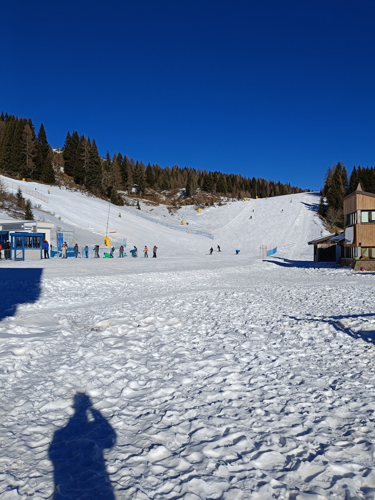 A winter scene captures a ski area with clear blue skies overhead. Snow-covered slopes are visible, dotted with skiers enjoying the terrain. In the foreground, a snow-covered ground is shown, while ski lifts and a reception area are positioned to the left.