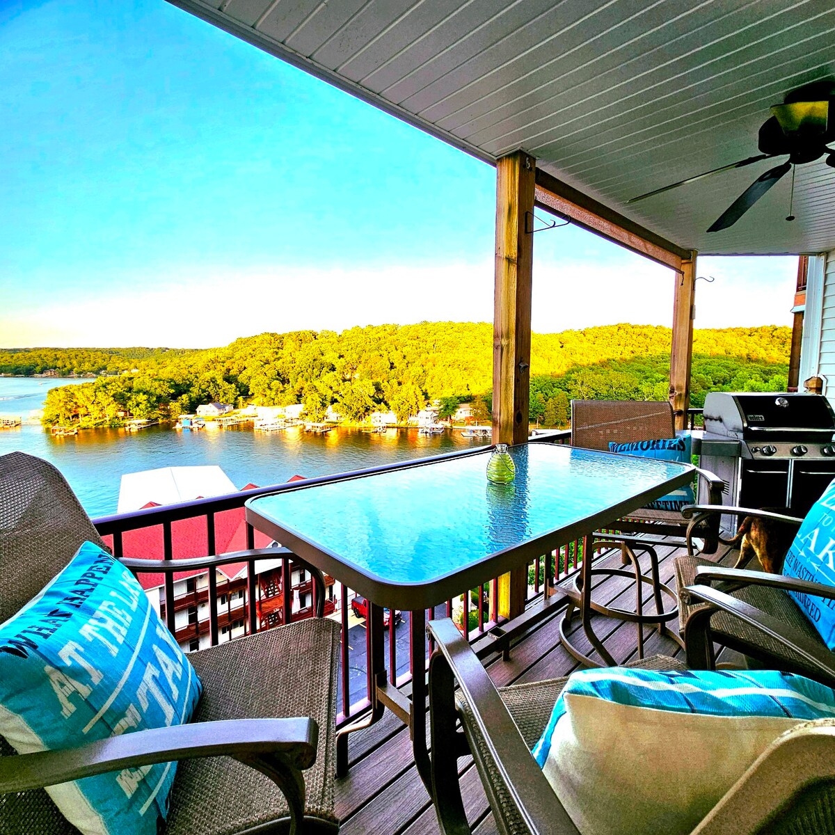 A covered balcony overlooks a serene lake, framed by lush greenery. A glass-top table is accompanied by comfortable seating with pillows. A grill is visible, adding to the outdoor dining experience. The tranquil water reflects the clear sky.
