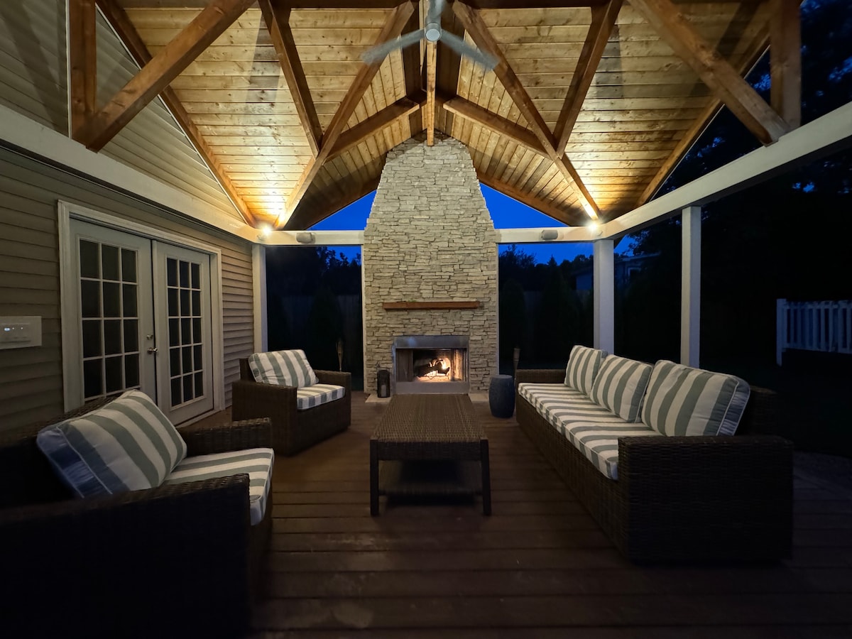 A covered outdoor seating area is showcased, featuring a stone fireplace that provides warmth. Comfortable seating with patterned cushions surrounds a central coffee table. The high wooden ceiling is accentuated by soft lighting, creating a relaxing ambiance as dusk approaches.