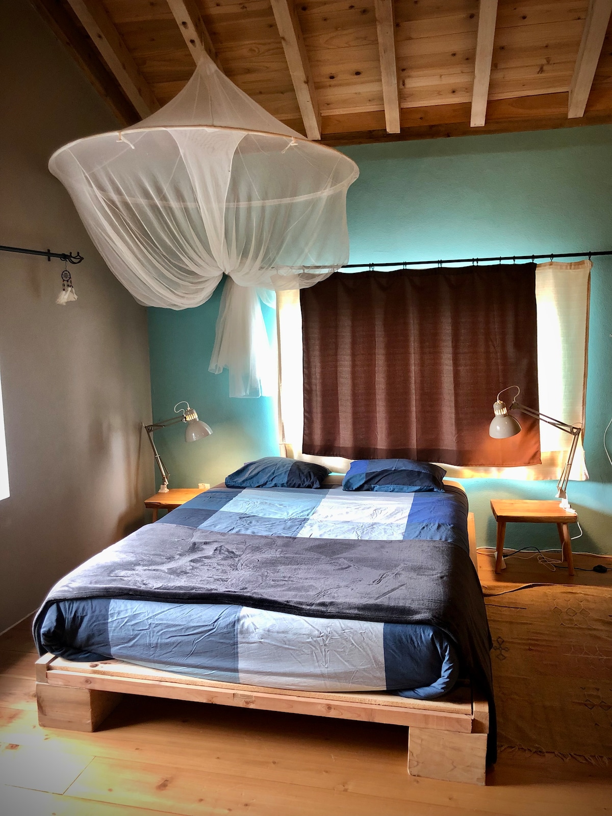 A spacious sleeping area features a wooden bed with a patchwork blanket in blue tones. Two bedside tables with lamps are situated on either side. Natural light enters through a window covered by dark curtains, while a mosquito net hangs from the ceiling.