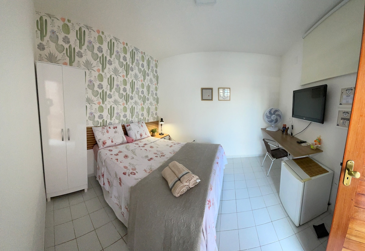 The suite features a spacious sleeping area with a floral bedding set on a large bed. A light-colored accent wall adorned with cactus patterns provides visual interest. A wooden desk with a chair, a compact refrigerator, and a television are present, alongside a wardrobe for storage.