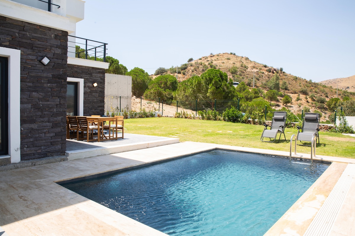 A modern outdoor area features a tranquil swimming pool surrounded by natural stone tiles. Sun loungers are positioned nearby, with a wooden dining table set under the sun. Lush greenery and hills can be seen in the background, enhancing the serene environment.