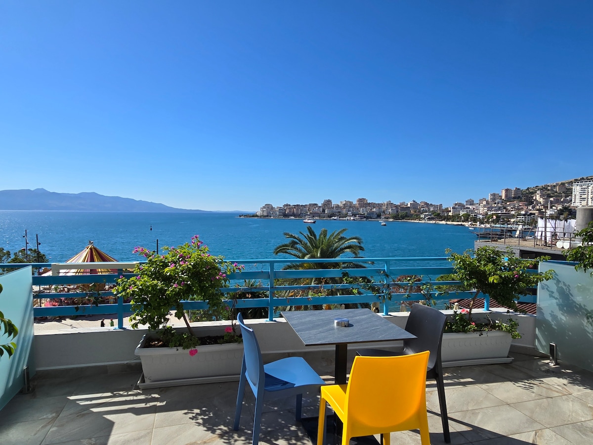 A balcony area is featured with a table surrounded by colorful chairs, overlooking a clear blue sea. Lush greenery is seen in planters alongside the railing, while the serene waterfront and distant hills create a calming backdrop.