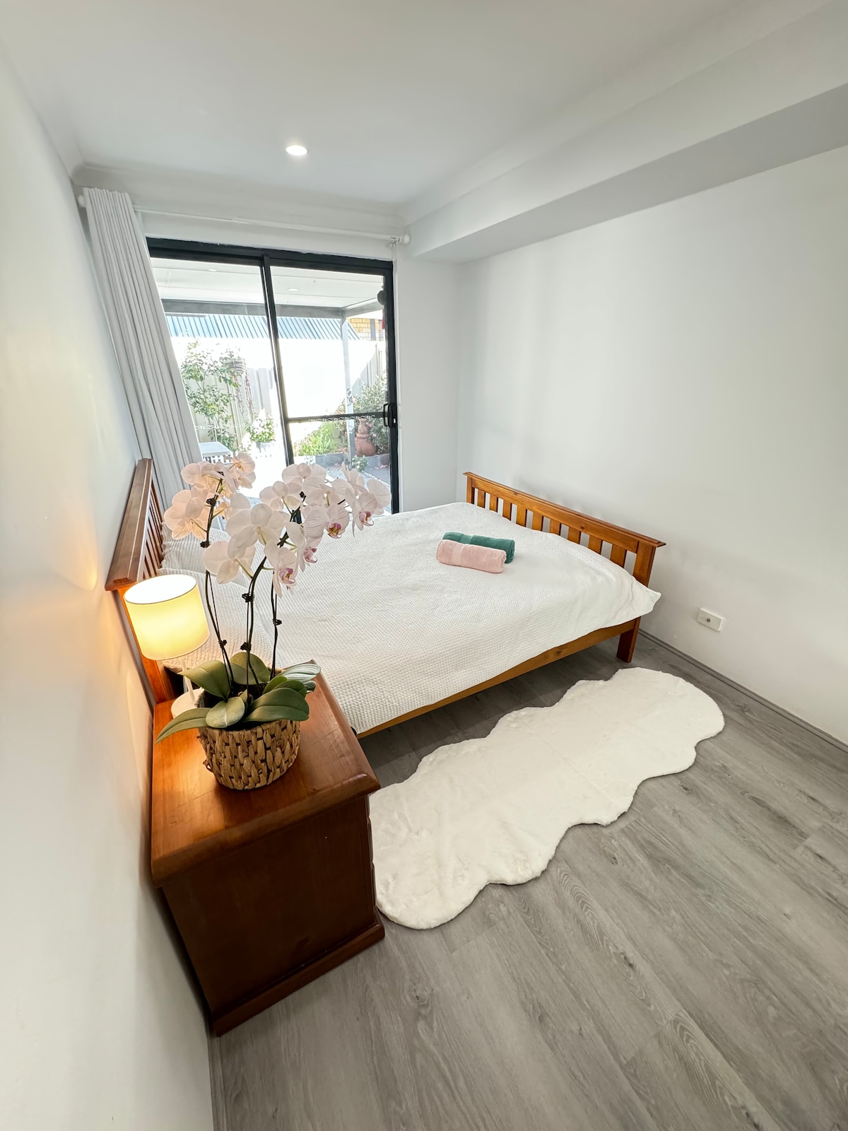 A well-lit bedroom features a spacious bed dressed in white linens. A bedside table holds a lamp and a decorative plant, while soft light filters through large glass doors leading to an outdoor area. A rug is placed on the wooden floor.