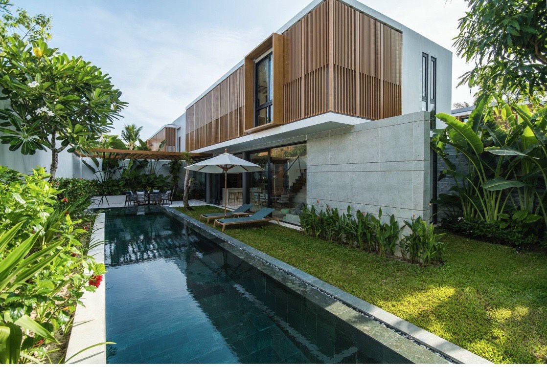 A modern home is surrounded by lush greenery, featuring a sleek swimming pool with dark water. Sun loungers are positioned beside the pool, and an umbrella provides shade. The architecture includes wooden slats and large windows that enhance the connection to the natural environment.